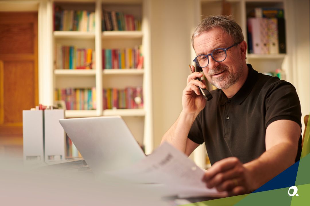 Man working from home office, speaking on the phone while reviewing financial documents on a laptop.