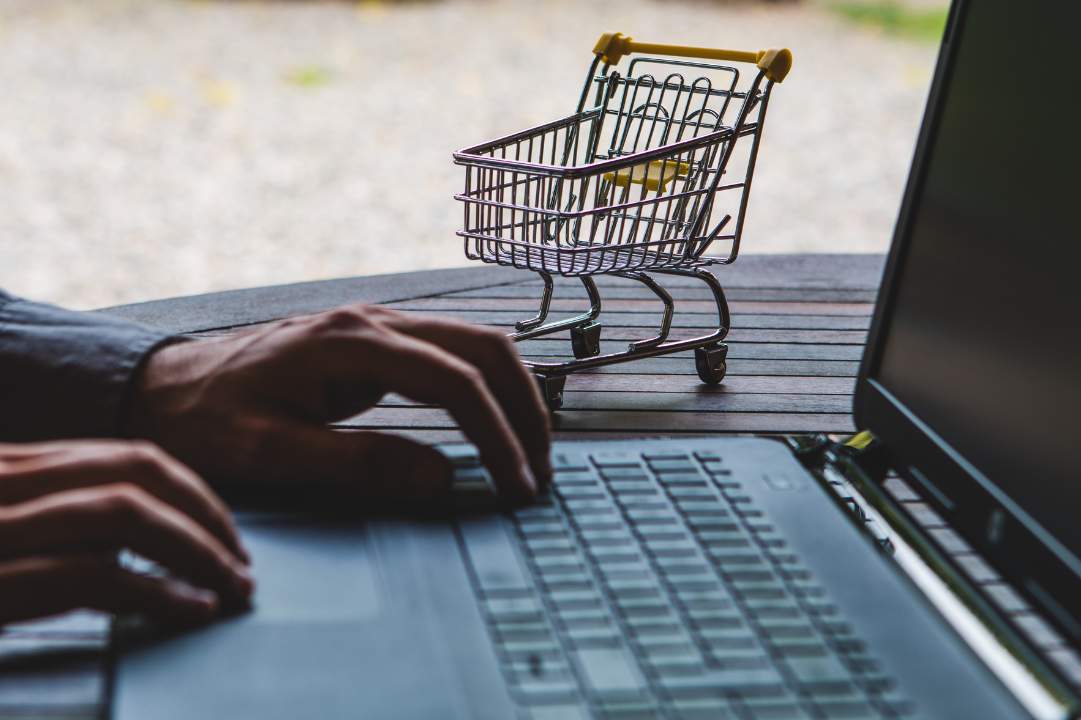 Photo of a hand using a laptop with a miniature shopping cart sitting next to it, signifying an ecommerce transaction