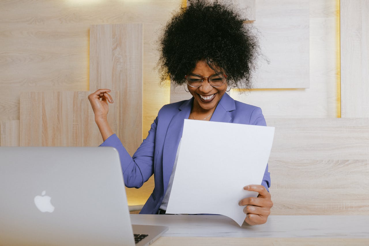 Woman holding a sheet of paper happy, sitting in front of a laptop